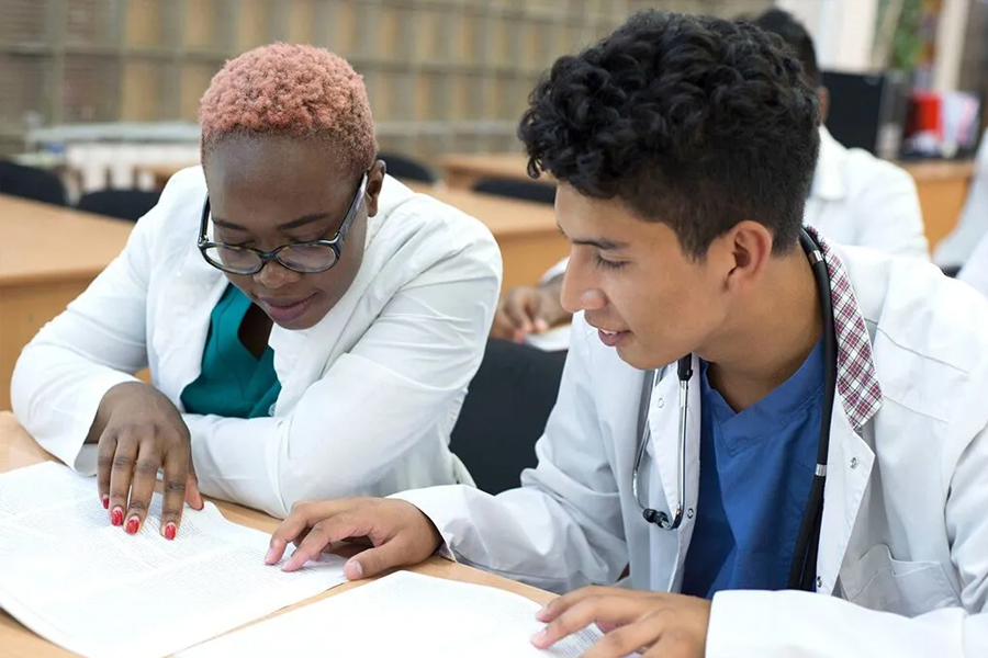 Two students in white coats read from a textbook together in a classroom